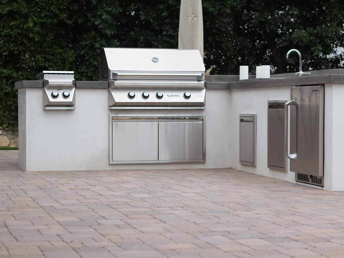 Long white outdoor kitchen island with built-in grill and stainless doors on a paver patio