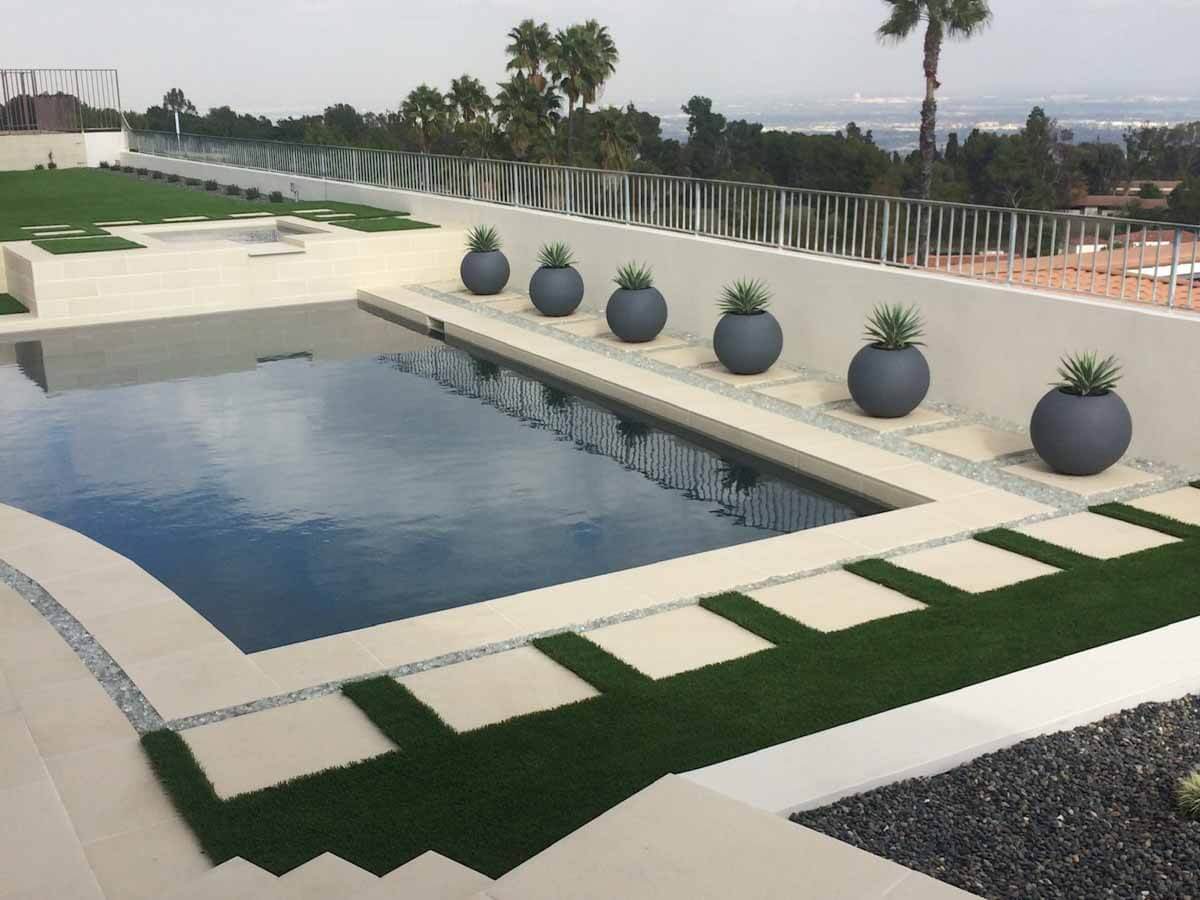 Modern pool with sphere planters along a glass fence, concrete pads and turf around it