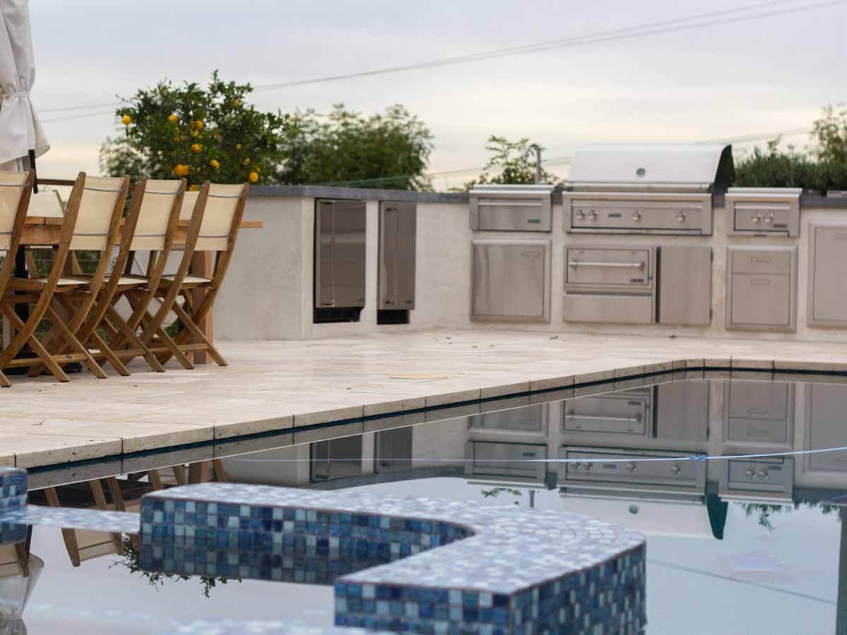 Glass tile spa and travertine pool deck with a full outdoor kitchen behind