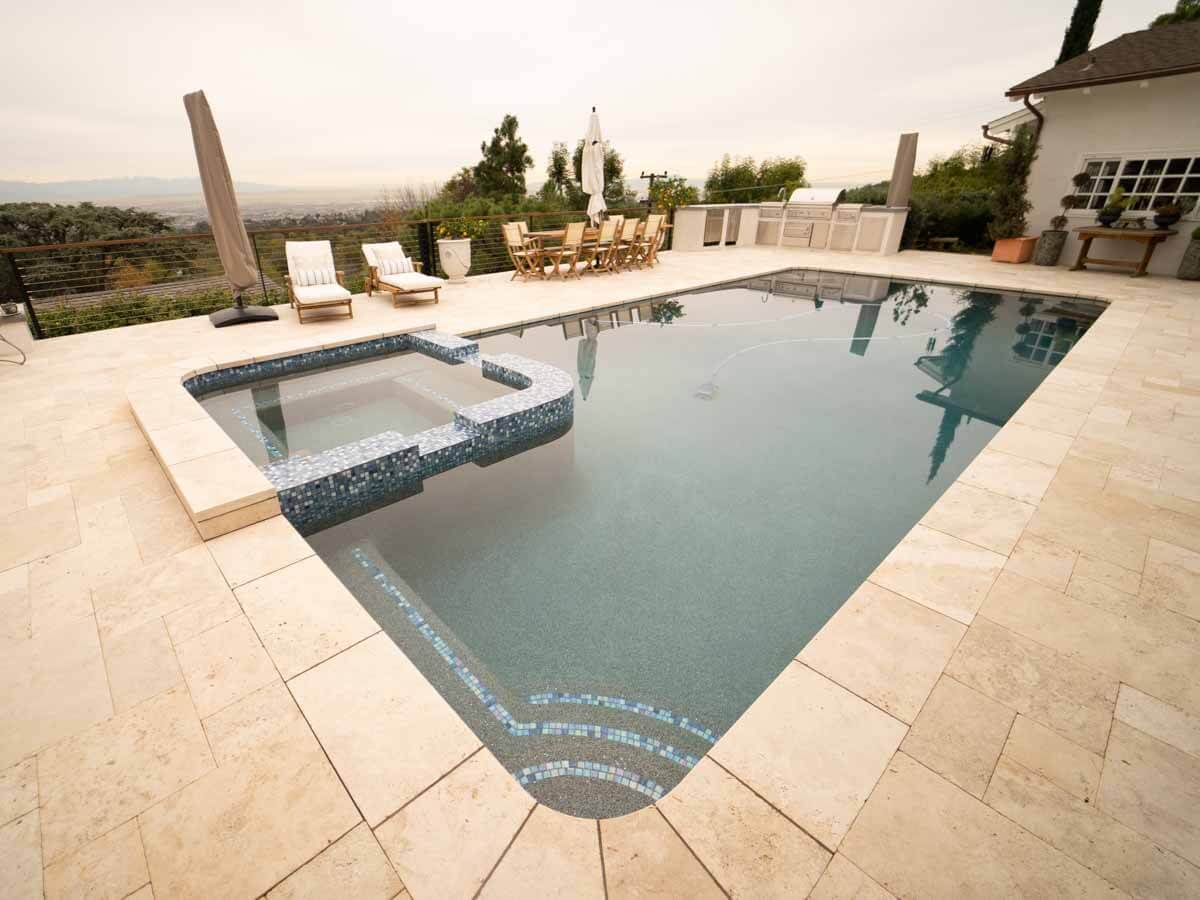 Raised spa clad in blue-gray tile spilling into the pool, travertine deck around it