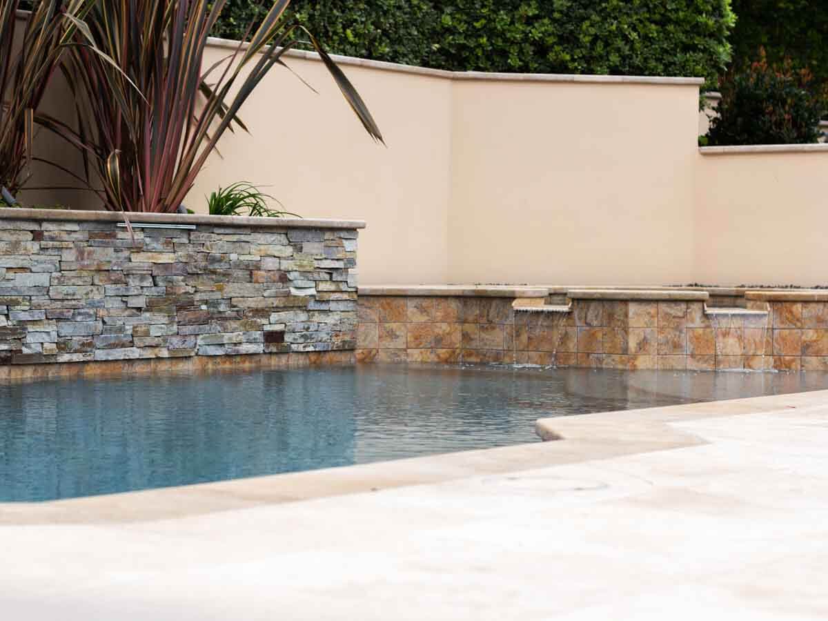 Stacked stone raised wall with spillways above a pool trimmed in travertine coping