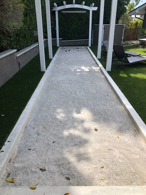 Bocce ball court with white concrete curbs beneath a white pergola, artificial turf and lounge chairs alongside