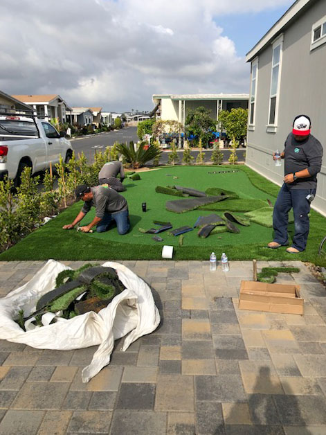 Roy's crew kneeling on a new green, fitting and trimming turf pieces beside a paver patio