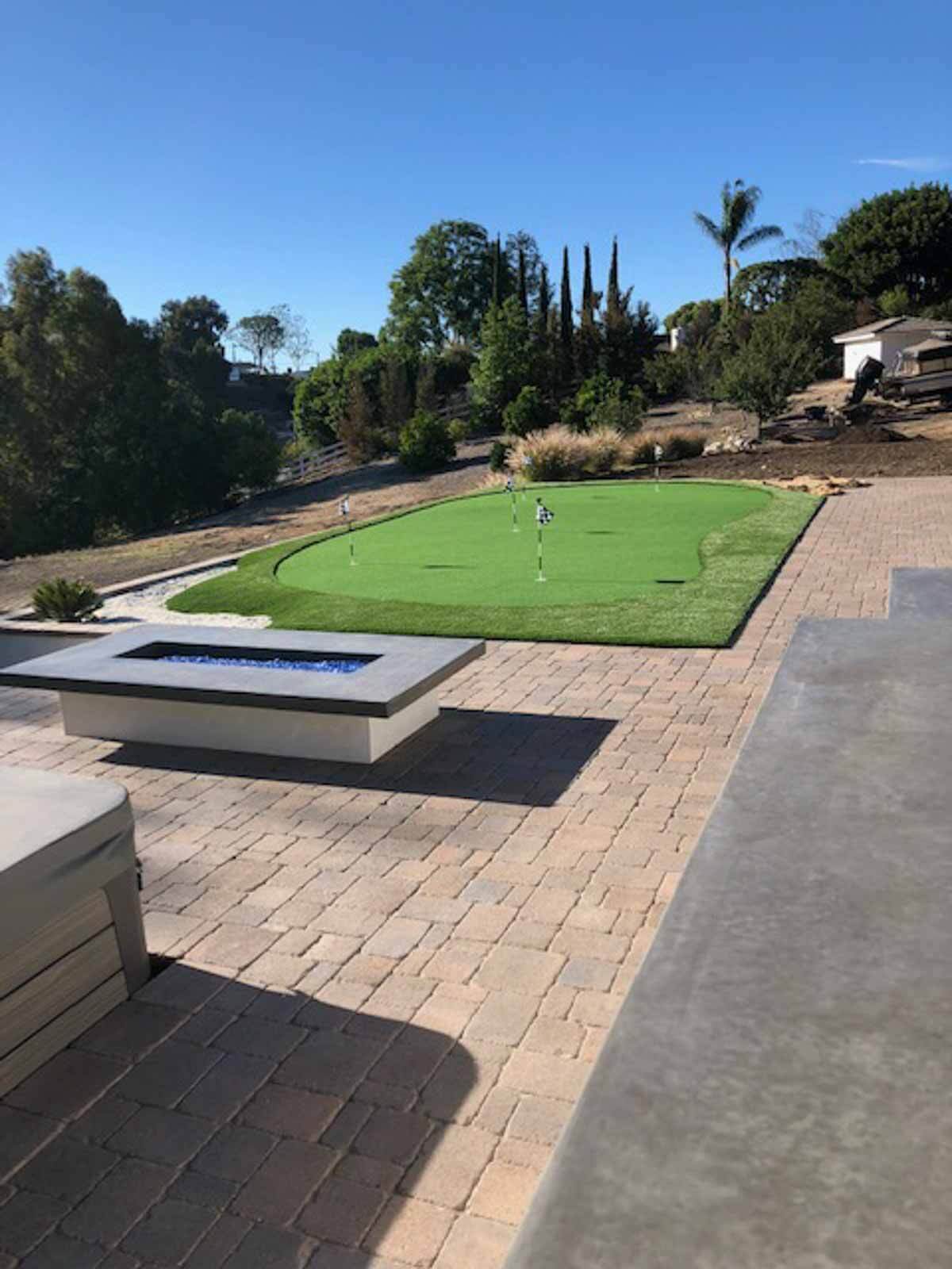 Putting green at the top of a paver terrace with a modern rectangular fire pit and a poured concrete walkway
